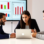 **

A diverse group of people, various ages and backgrounds, gathered around a table looking at charts and graphs on a laptop screen. The setting is a bright, modern co-working space in Madrid. Focus on expressions of understanding and collaboration. Some are pointing at the screen with interest. The overall feel is positive and empowering, representing learning about investment and financial planning. In the background, a whiteboard with the words "Metas Financieras" (Financial Goals) and "Interés Compuesto" (Compound Interest).

**